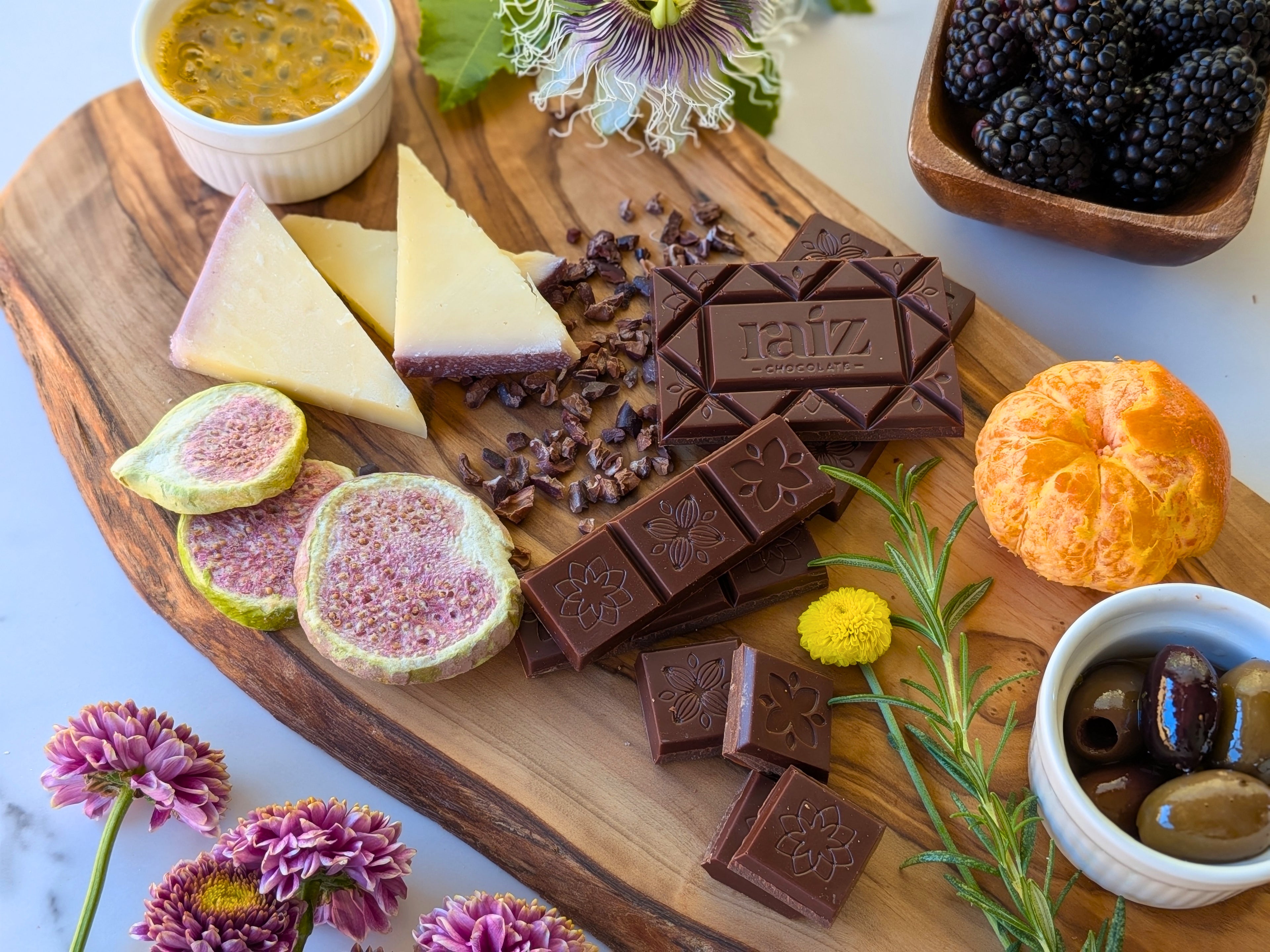 Wooden charcuterie board with assorted cheeses, chocolates, fruits, and olives on a white surface.